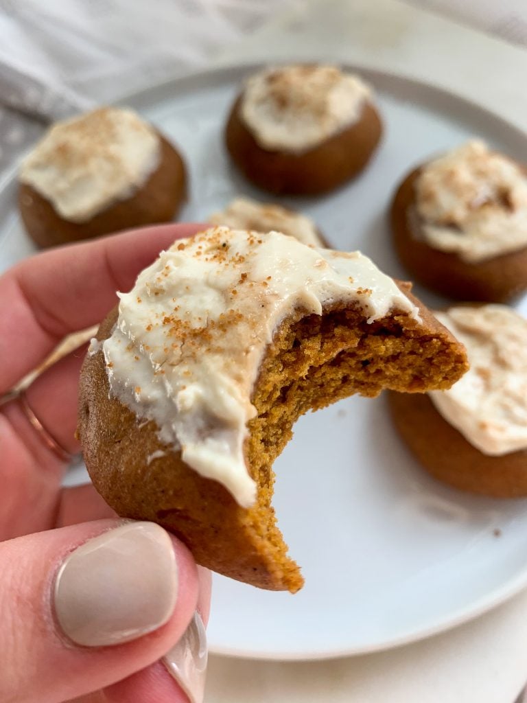 a hand holding a whole wheat pumpkin cookie with a maple cream cheese frosting over a white plate