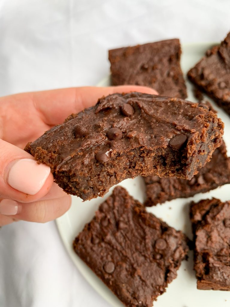 a hand holding a fudgy chickpea brownie over a white plate of more brownies