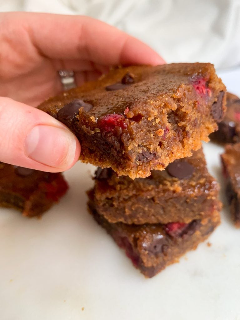 a hand holding a raspberry chocolate chip blondie over a white background