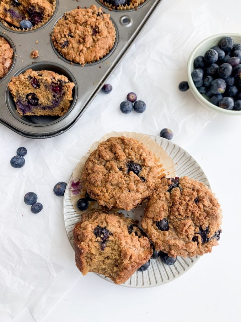 a plate of muffins, bowl of blueberries, and muffin tin on a white background