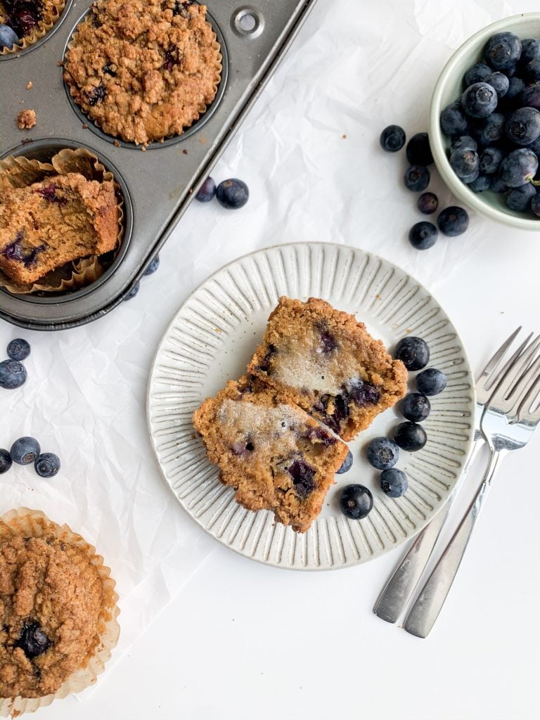 a blueberry muffin on a gray plate, a bowl of blueberries, and a muffin tin on a white background