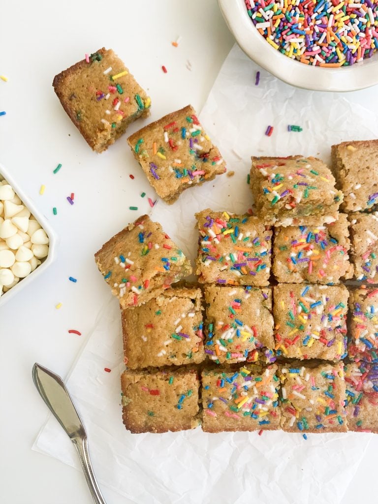 white background with blondies and a bowl of sprinkles and white chocolate chips