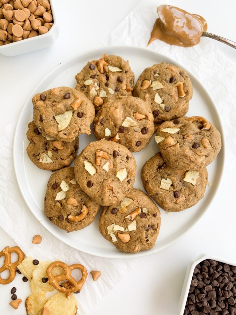 white plate of cookies surrounded by chocolate chips, pretzels, potato chips, and peanut butter