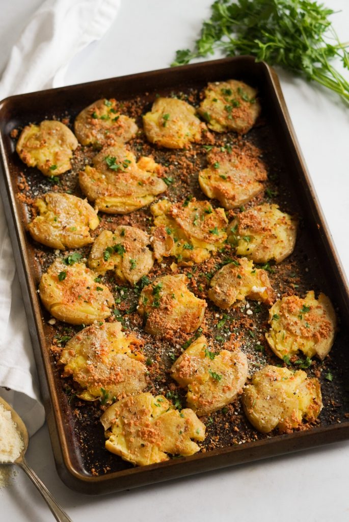 crispy smashed potatoes on a baking tray on a white background with parsley and a spoonful of parmesan cheese on the side