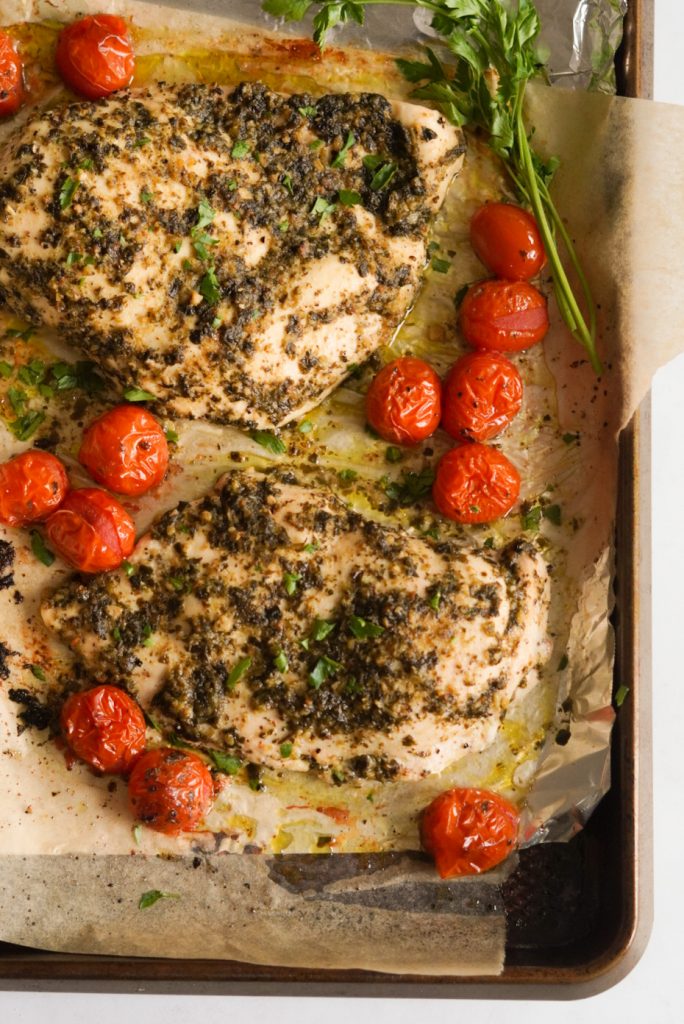 easy pesto chicken, tomatoes, and fresh parsley on a parchment paper lined baking sheet