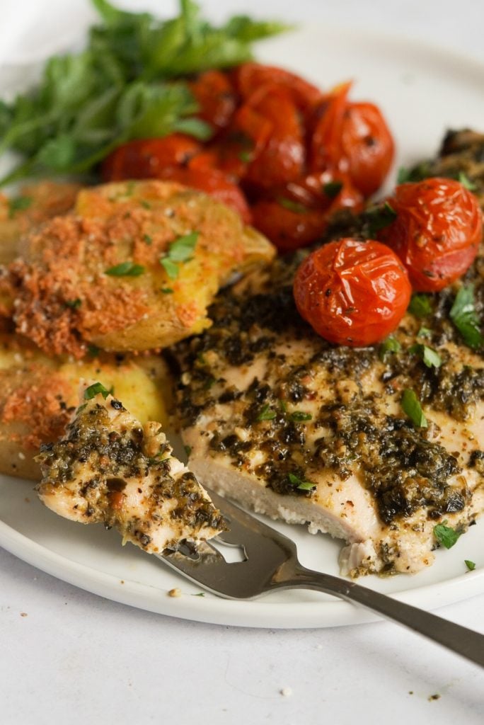 pesto baked chicken with a piece cut off on a fork, grape tomatoes, and smashed potatoes on a plate