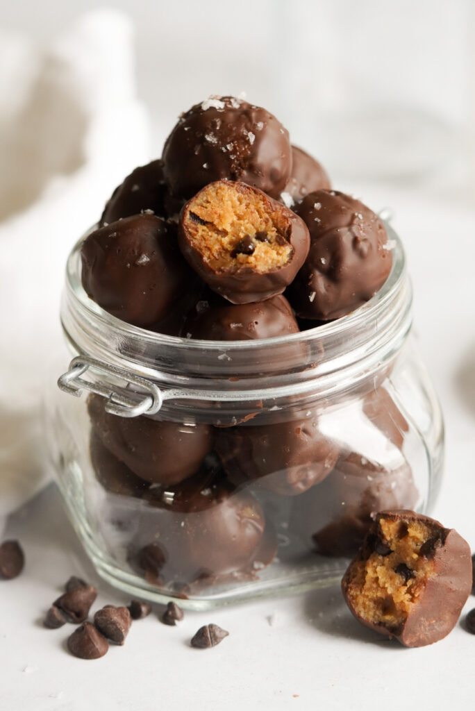 chocolate coated cookie dough balls in a jar on a white background, one has a bite taken out of it