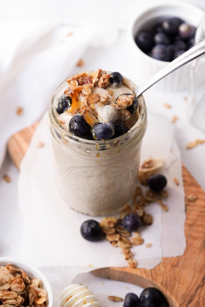 blended overnight oats in a jar topped with blueberries, granola, and honey on a wooden platter surrounded by a side of granola, a side of blueberries, and a honey stick