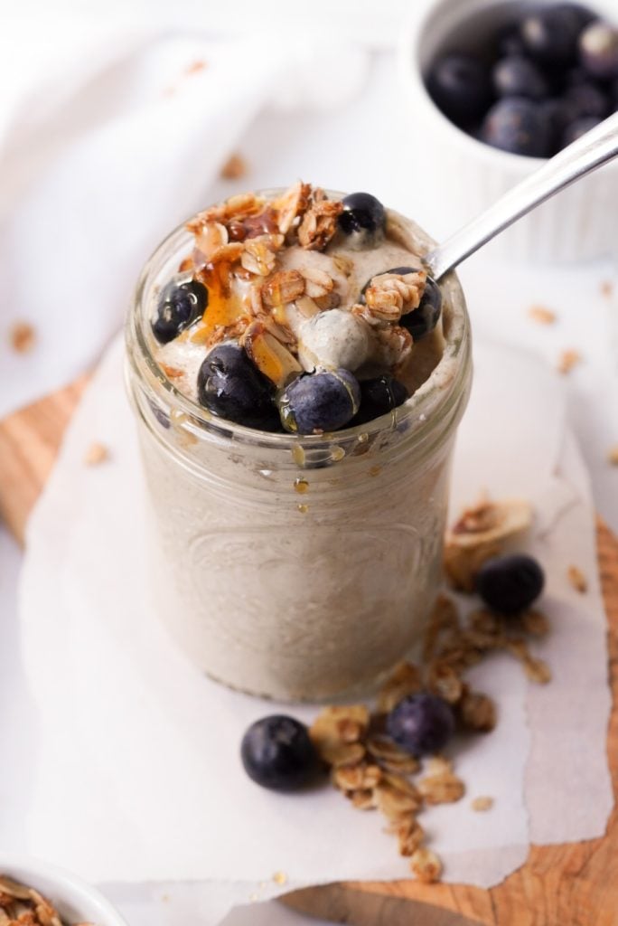 blended overnight oats in a jar topped with granola, honey, and blueberries on a wooden tray with parchment paper