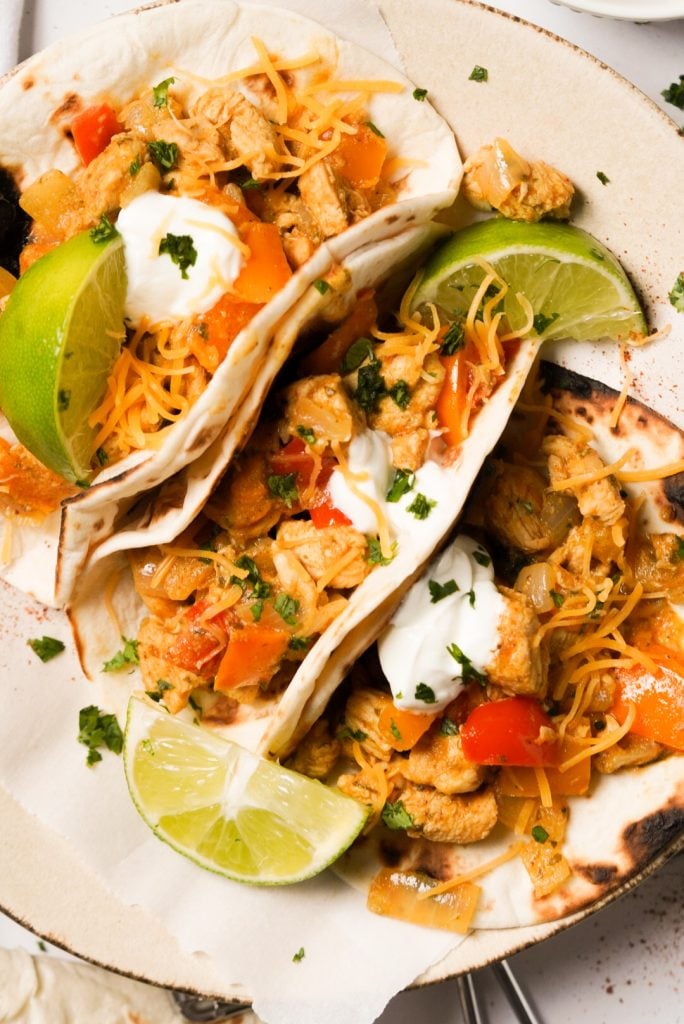 Overhead shot of 3 assembled chicken fajita tacos on a white plate with lime wedges scattered around.