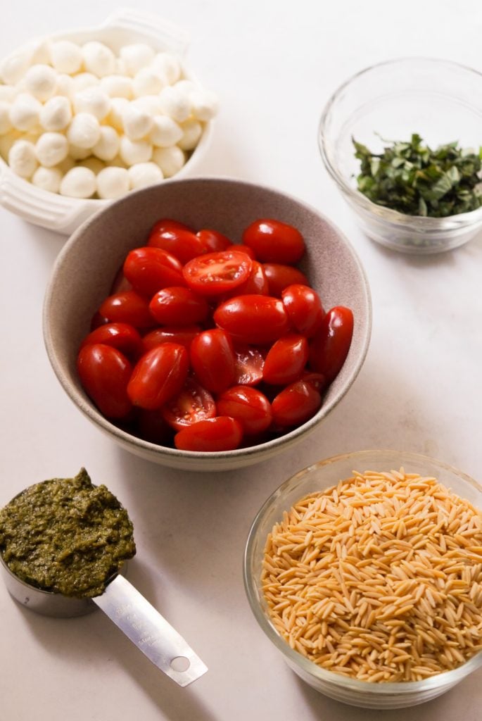Ingredients needed to make orzo pasta salad with pesto measured out into bowls on a white table.