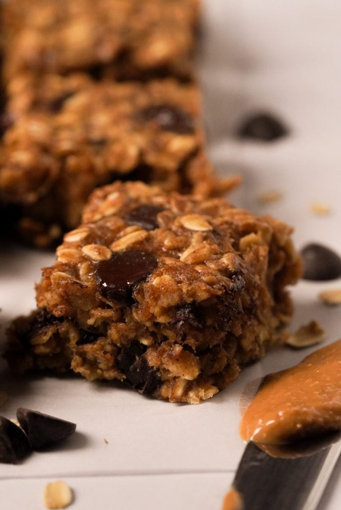 Side-on closeup shot of banana oatmeal bar with chocolate chips next to a butter knife loaded with peanut butter.