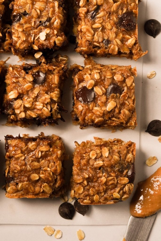 Overhead shot of banana peanut butter oat bars that have been sliced into squares for serving on a piece of parchment with rolled oats and chocolate chips sprinkled around.