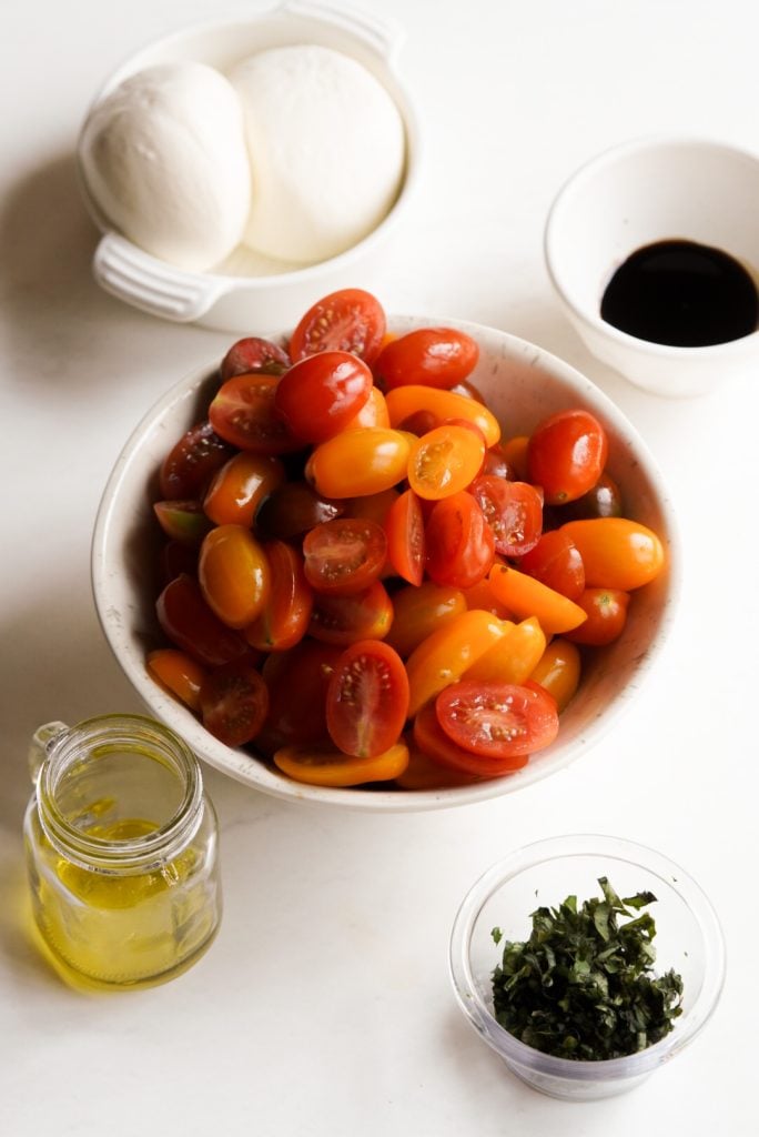 ingredients needed to make burrata caprese with cherry tomatoes measured out into bowls on a white table.