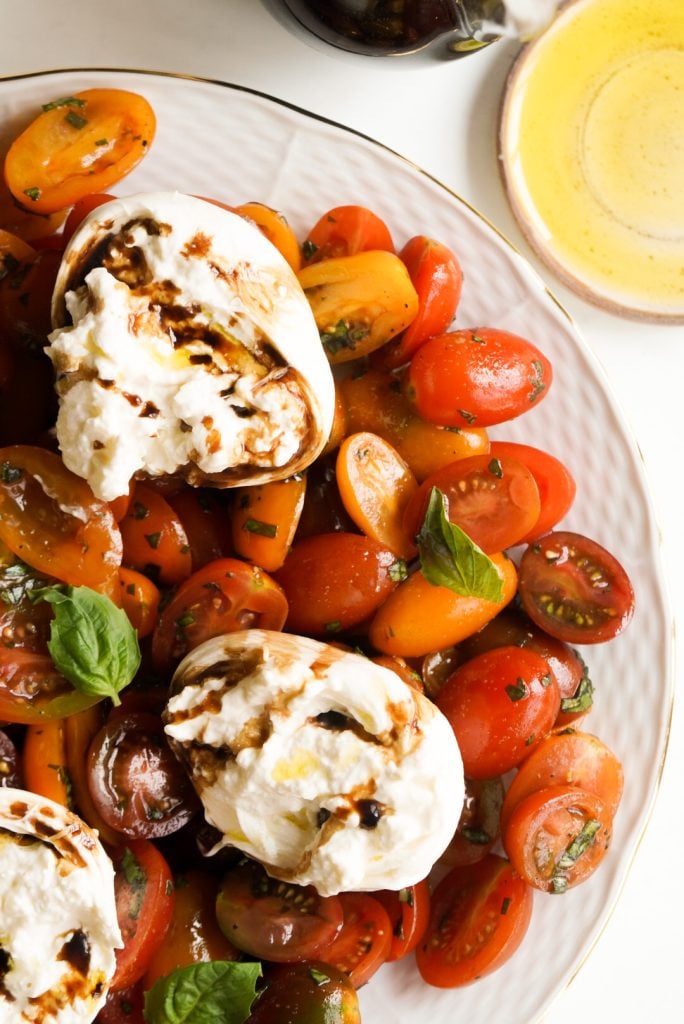 overhead hero shot of a white serving platter of burrata caprese on a white table next to a shallow bowl of evoo.