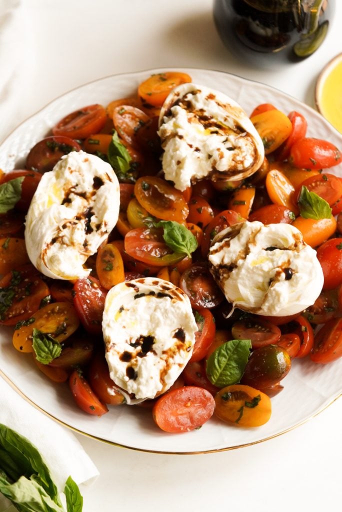 table with a serving platter of fresh tomato caprese salad with burrata cheese next to a cruet of balsamic vinegar and a bunch of fresh basil on a white table.