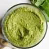 blended cashew pesto in a clear bowl with fresh basil leaves, raw cashews, and fresh garlic scattered about on the table.