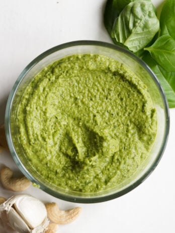 blended cashew pesto in a clear bowl with fresh basil leaves, raw cashews, and fresh garlic scattered about on the table.