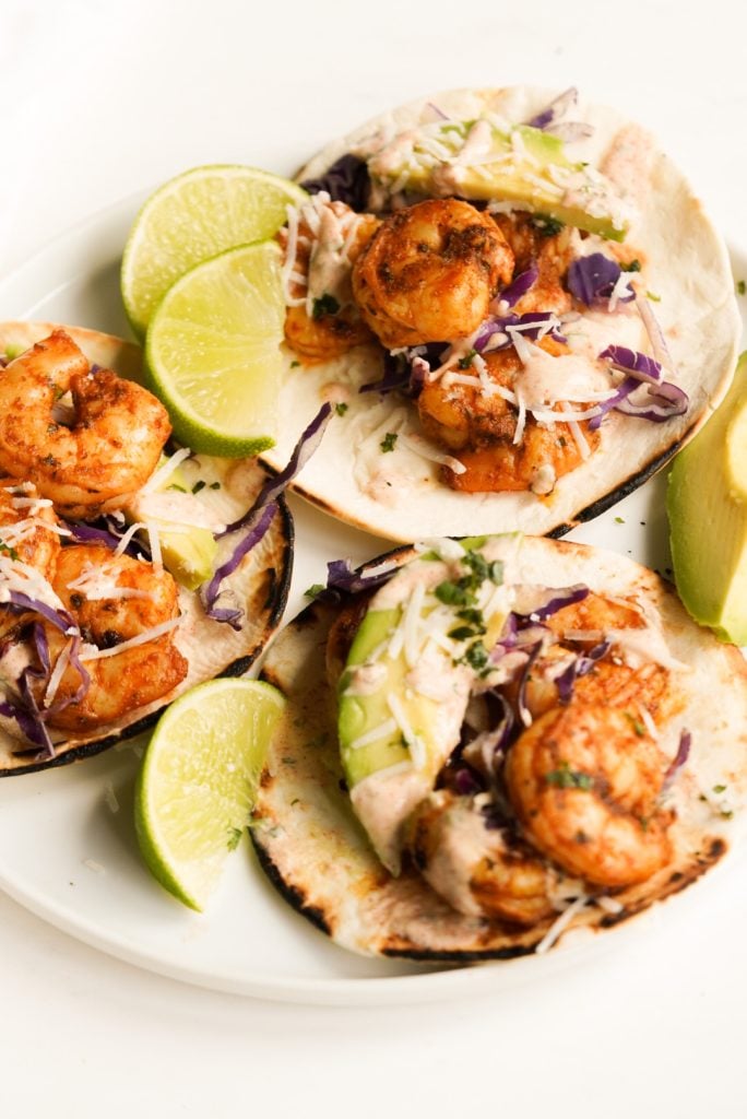 45 degree hero shot of a serving platter with 3 assembled baja shrimp tacos served with extra wedges of lime and avocado.