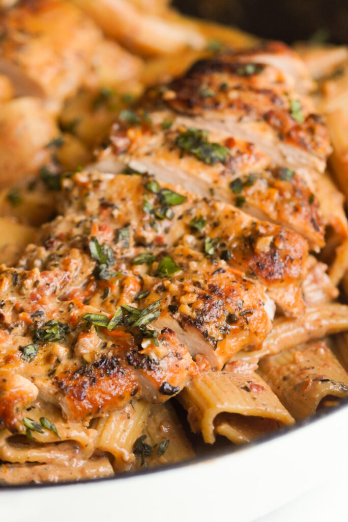 sliced seasoned chicken breast on top of a skillet filled with creamy sundried tomato pasta.