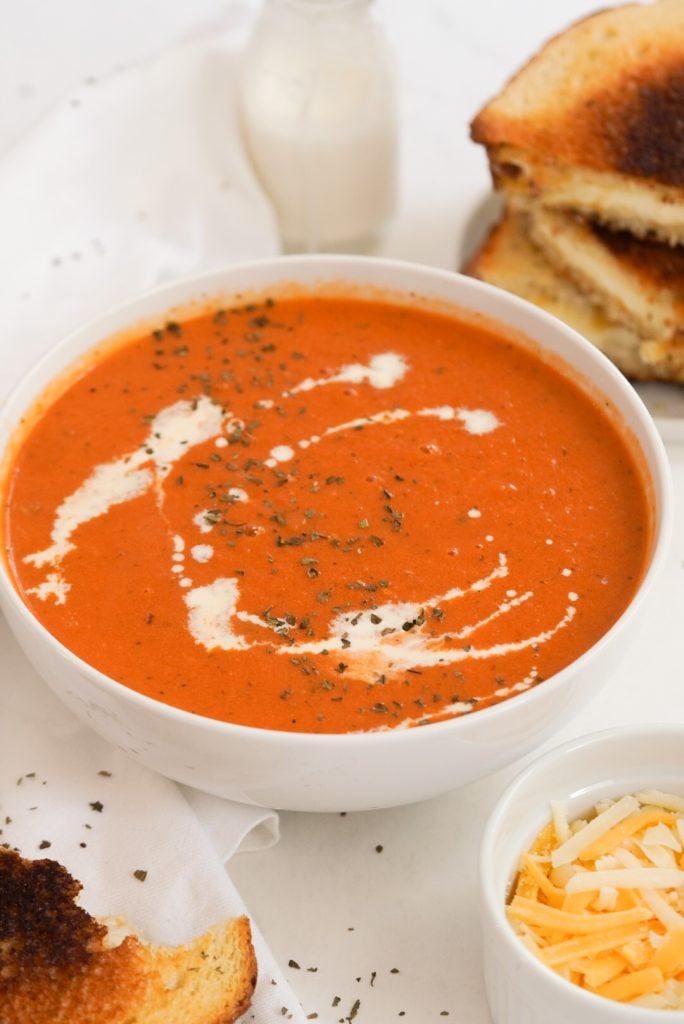 45 degree angle shot of a bowl of tomato soup with a stack of grilled cheese sandwiches.