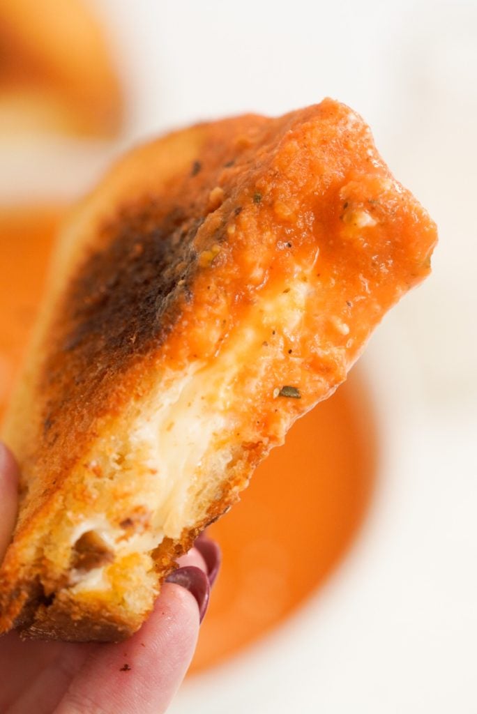 closeup shot of a hand holding a piece of grilled cheese that has been dunked in tomato soup.