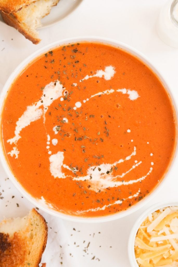 white bowl of tomato soup drizzled with cream and served with grilled cheese on the side.