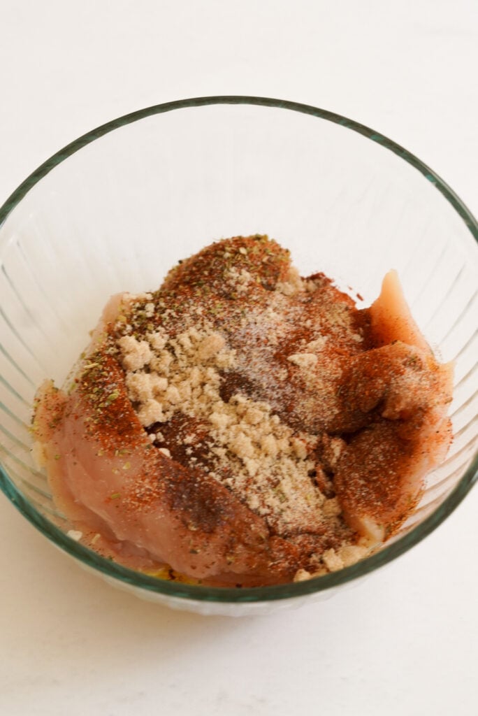 process shot of raw chicken tenderloins in a mixing bowl with the spices and seasonings on top.