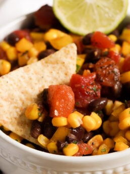white ribbed bowl with a tortilla chip taking a scoop of corn black bean salsa with a halved lime.