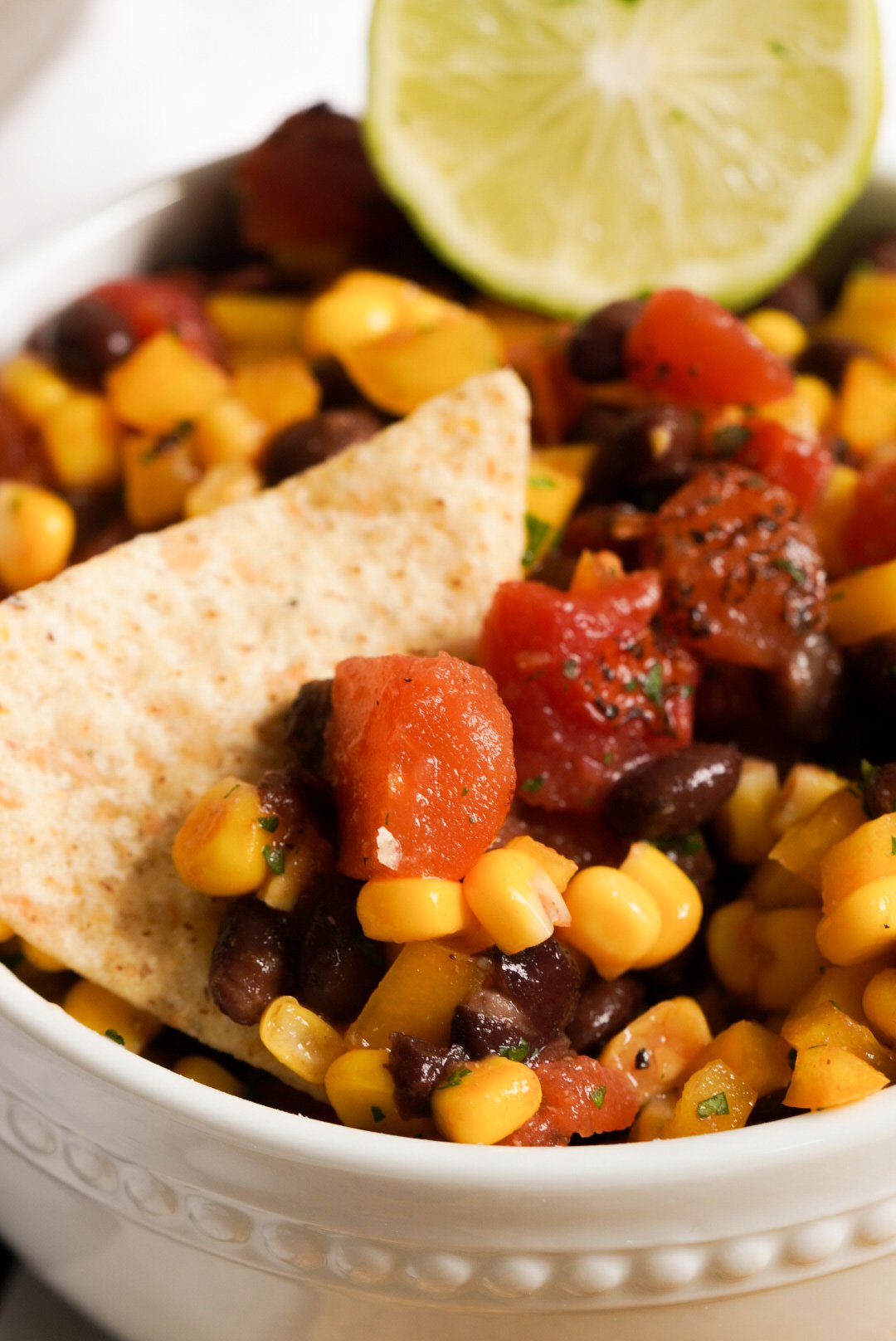 white ribbed bowl with a tortilla chip taking a scoop of corn black bean salsa with a halved lime.