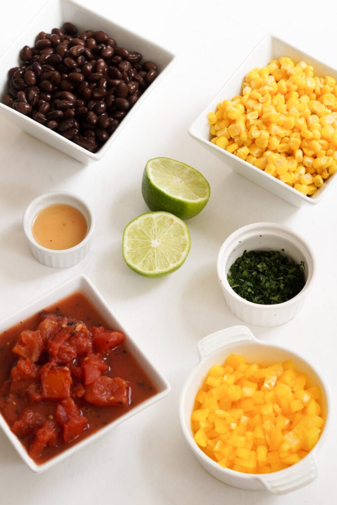 flat lay shot of the ingredients needed to make corn and black bean salsa measured out into bowls on a white table.