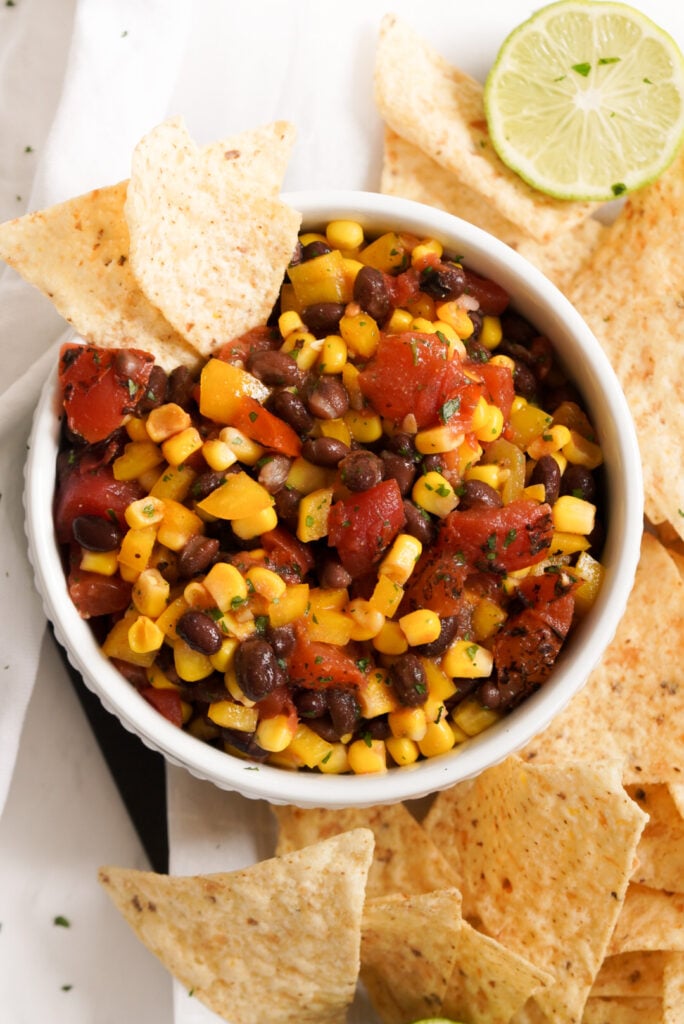 overhead flat lay shot of a white bowl of easy corn and black bean salsa with 2 tortilla chips sticking out of the bowl and a bunch of tortilla chips and a halved lime spread around the bowl.