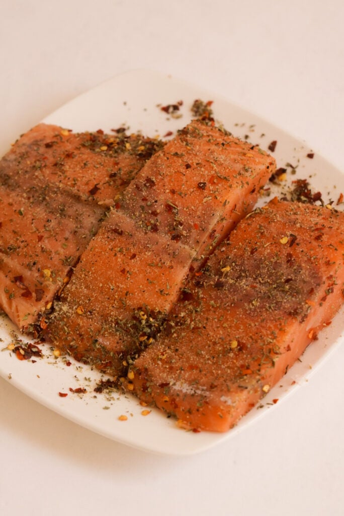 3 salmon fillets on a square white plate with seasoning added.