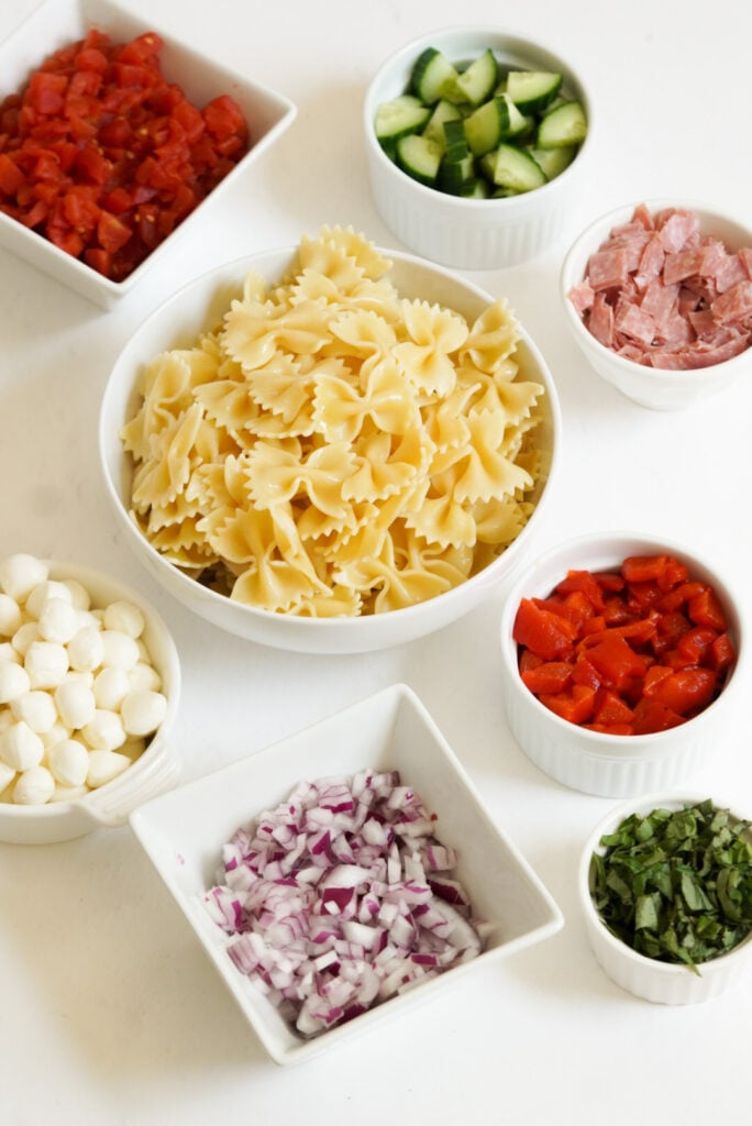 ingredients for italian bow tie pasta salad measured out into bowls on a white table.
