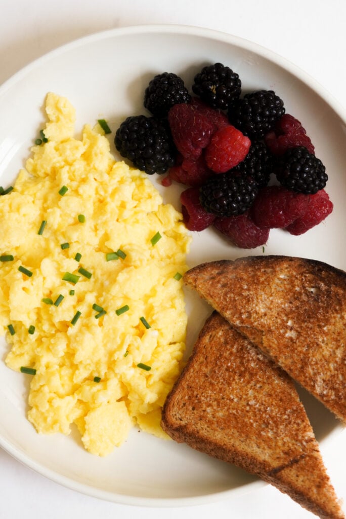 flat lay shot of a white plate with cottage cheese scrambled eggs topped with chives on the left, fresh raspberries and blackberries in the top right quadrant, and a halved slice of whole wheat toast stacked in the bottom right. 