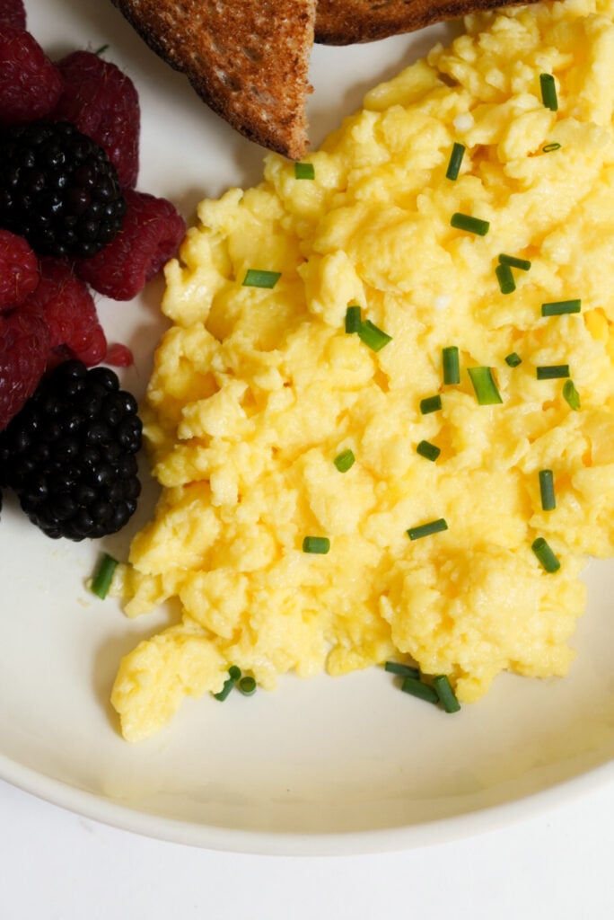 creamy scrambled eggs with cottage cheese topped with chopped chives on a white plate with fresh berries and toast. 