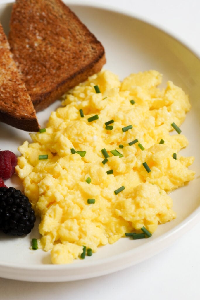 white plate with focus on cottage cheese scrambled eggs garnished with chives, plus a few slices of wheat toast and fresh berries to the side. 