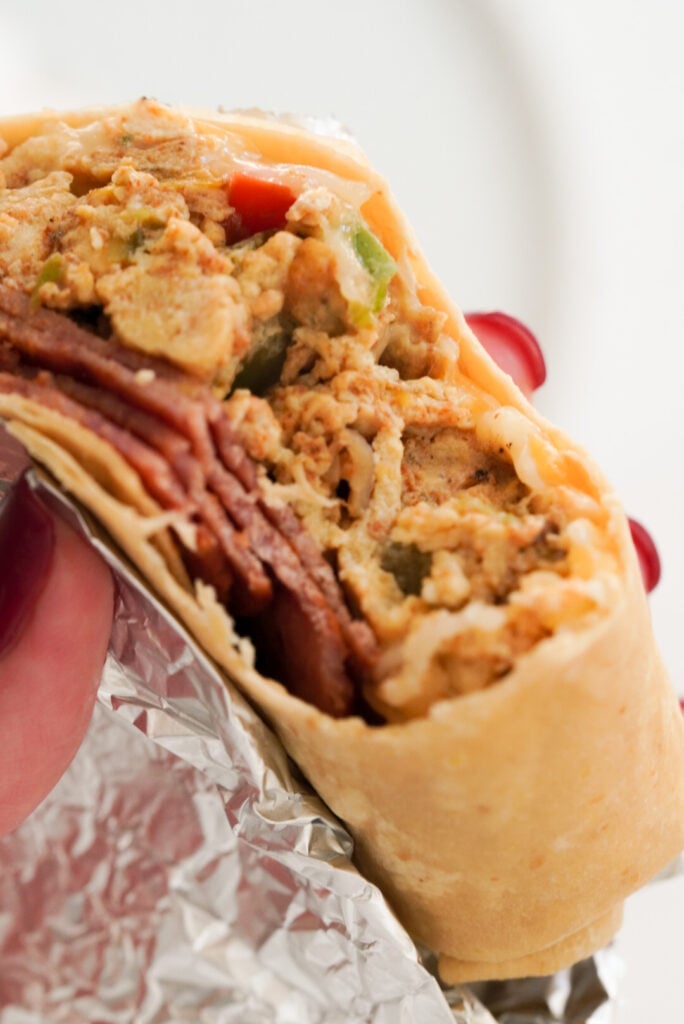 closeup shot of a bacon and egg breakfast burrito with a bite taken out and the foil slightly unwrapped being held by a woman's hand with red nail polish.