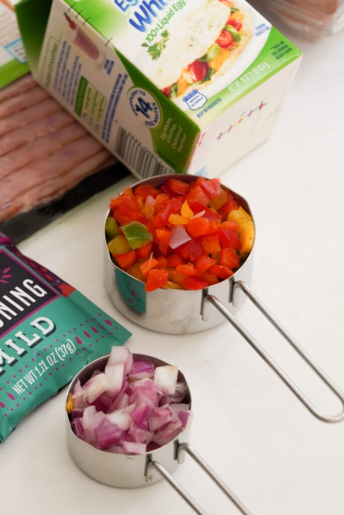 silver measuring cups with diced onions and diced peppers on a white table next to a carton of egg whites.