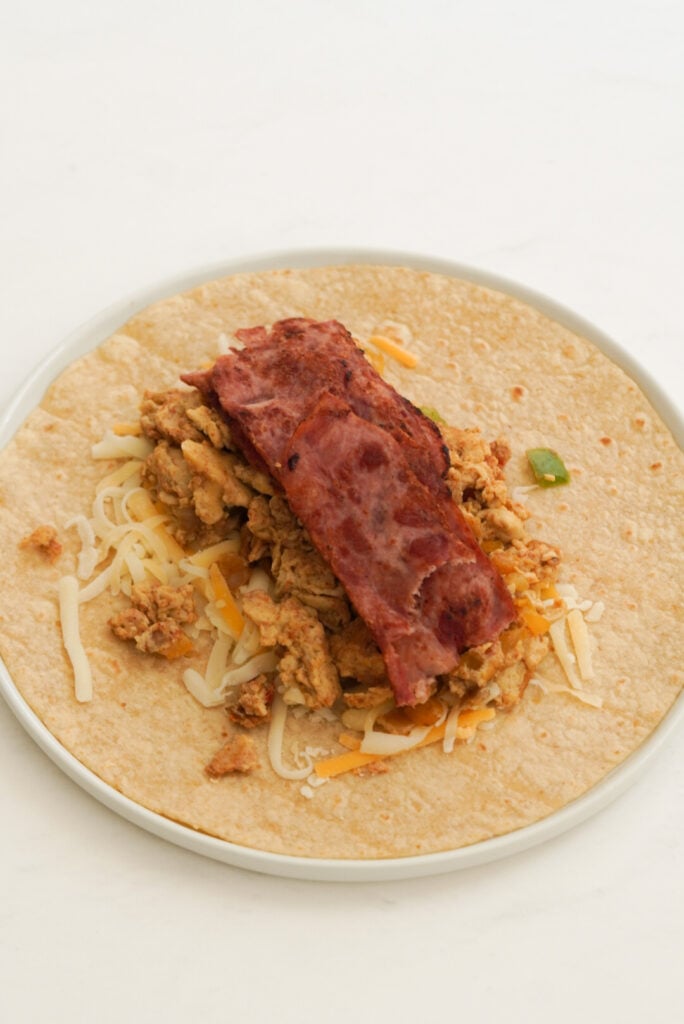 open faced tortilla with veggie egg scramble, cheese, and turkey bacon layered in the middle for wrapping into a burrito.