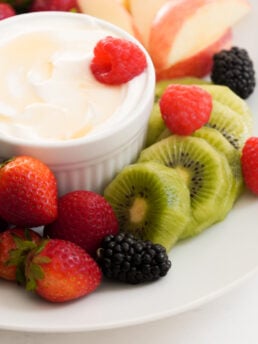 sliced apples, sliced peeled kiwi, whole raspberries, blackberries, and strawberries on a round white plate with a ramekin of fruit dip made with greek yogurt and honey.