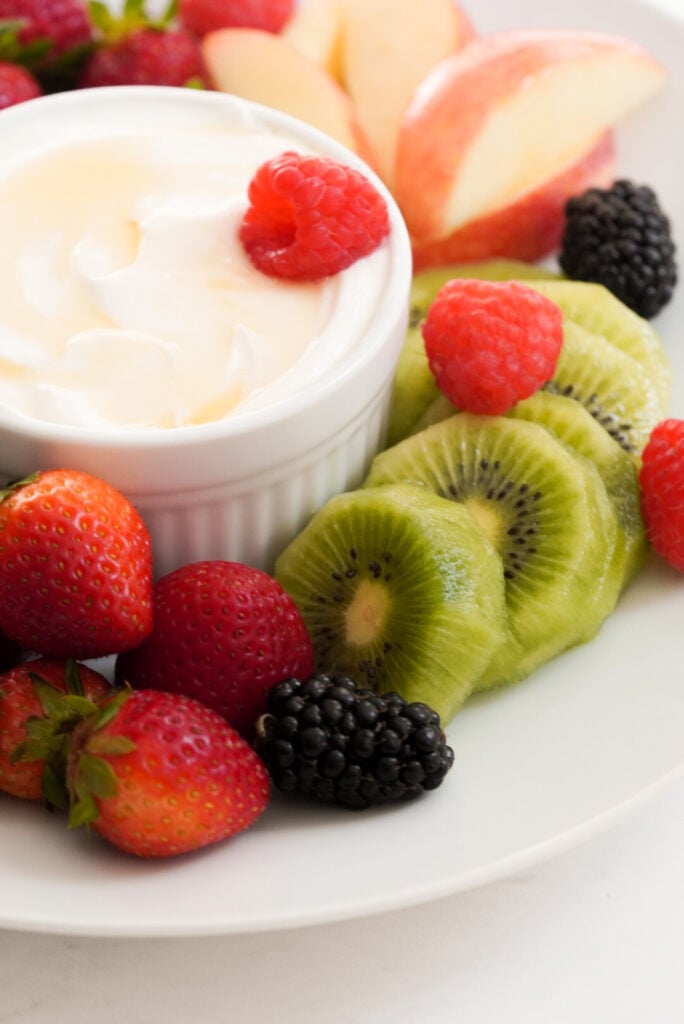 sliced apples, sliced peeled kiwi, whole raspberries, blackberries, and strawberries on a round white plate with a ramekin of fruit dip made with greek yogurt and honey.