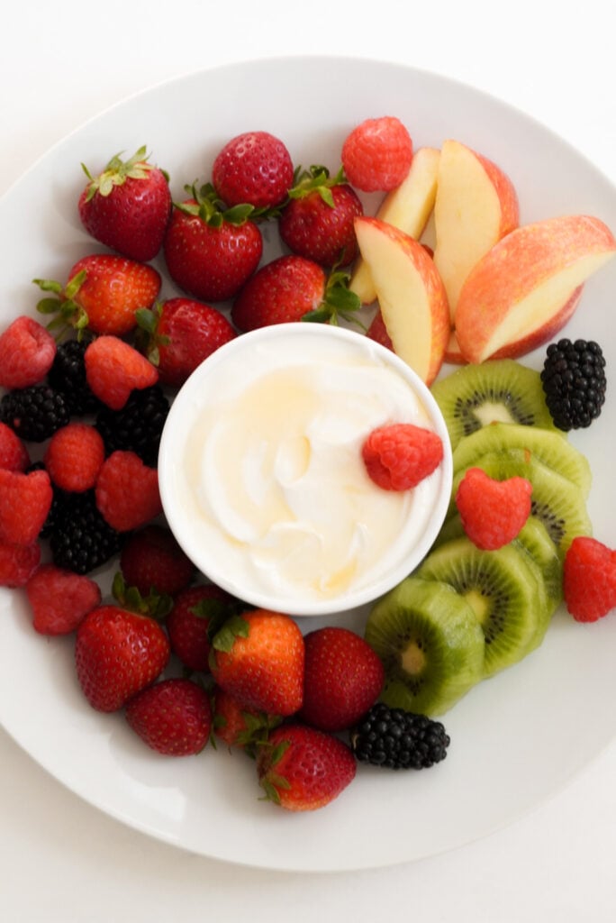 small white bowl of fruit dip made with greek yogurt and honey on a white plate with colorful fresh fruit.