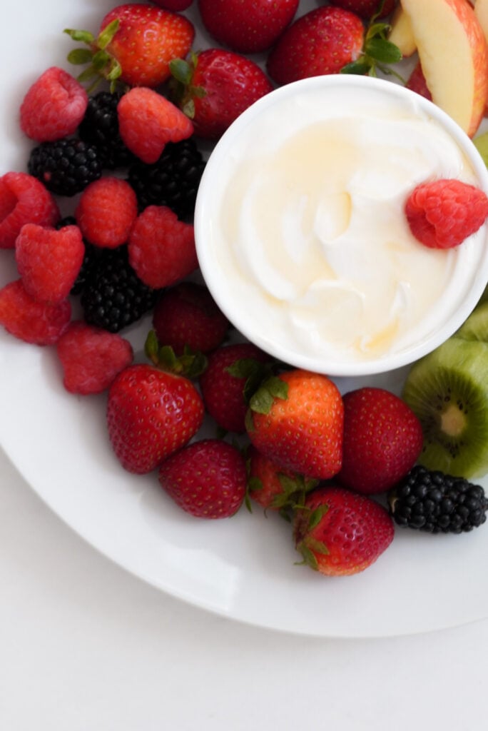 flat lay shot of greek yogurt dip set up for dipping fruit into; whole berries surround the ramekin to the left and beneath it.