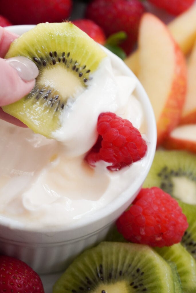 hand dipping a round slice of kiwi into a bowl of greek yogurt fruit dip in a white ramekin with a red raspberry stuck to the dipped end.