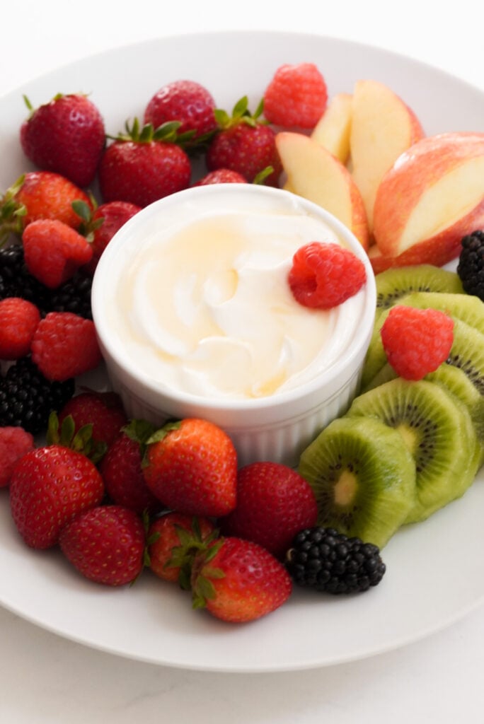 white grazing platter set up with a white ramekin of greek yogurt honey fruit dip in the middle and fresh fruit surrounding it on a white table.