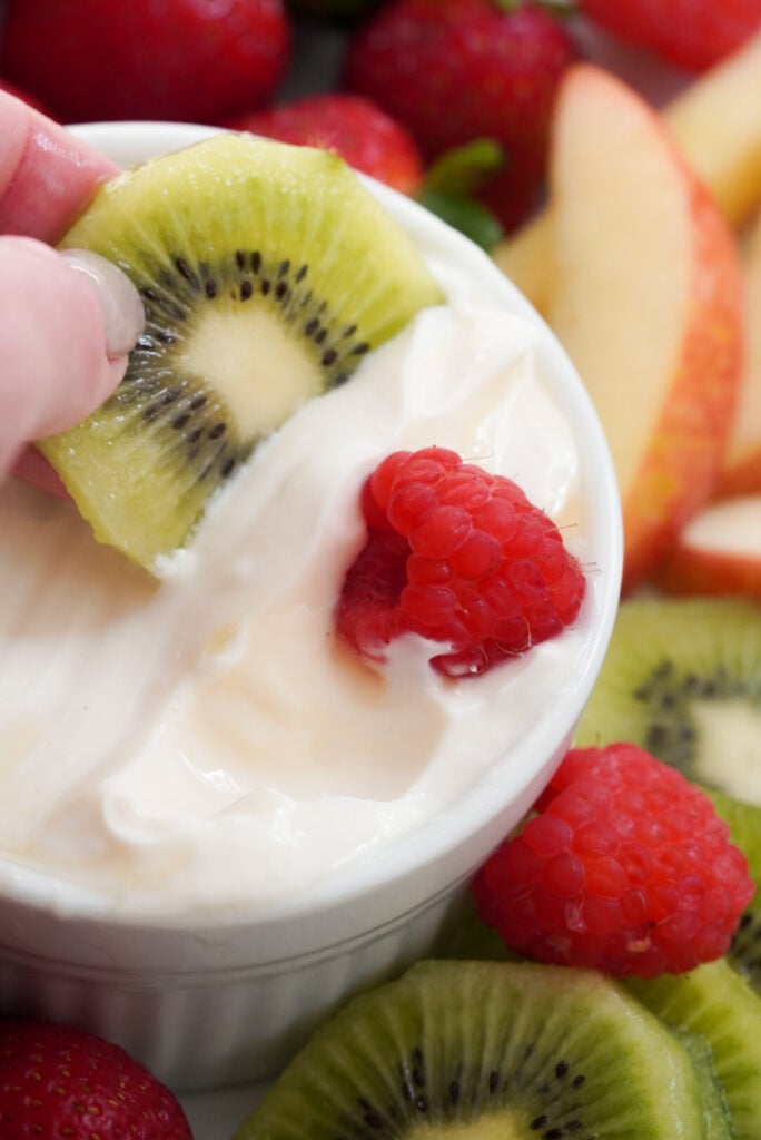 hand dipping a slice of kiwi into a round white ramekin filled with healthy greek yogurt and honey fruit dip on a tray surrounded with more fresh fruit.