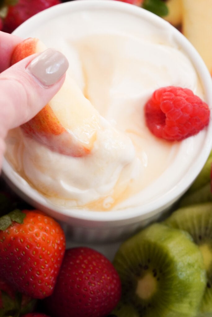 overhead shot of an apple slice being dipped into a ramekin of healthy greek yogurt fruit dip with a raspberry on the right side of the ramekin and whole strawberries and kiwi slices surrounding the bowl.