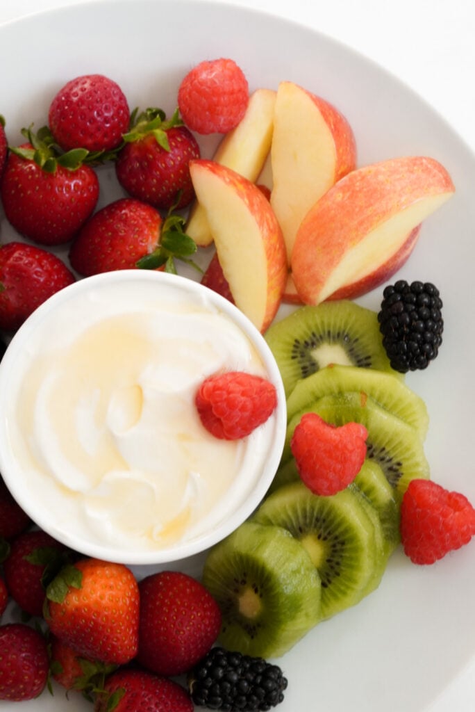 overhead shot of yogurt fruit dip that has been drizzled with honey on a round white tray surrounded with a rainbow of fresh fruit.