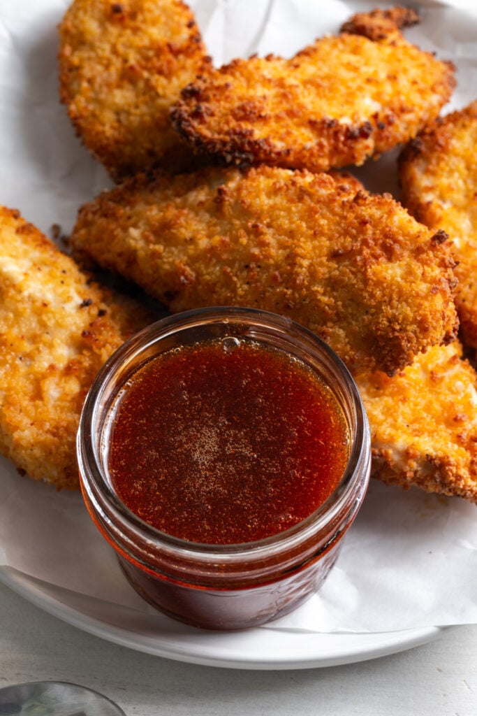 naked spicy chicken tenders on a plate with a squat short mason jar filled with a deep red hot honey sauce.